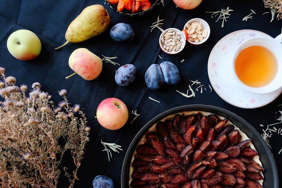 Flat lay of colorful fruits, plum tart, and herbal tea on a dark table setting.