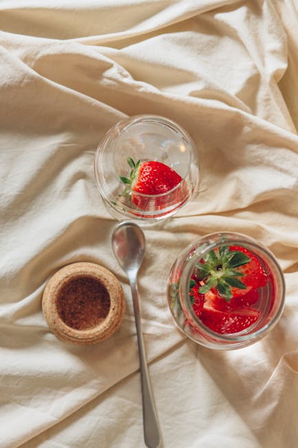 Top view of fresh strawberries in glass jars with a spoon on a textured fabric background.