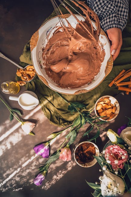 Overhead view of chocolate batter surrounded by ingredients and flowers on a rustic table.