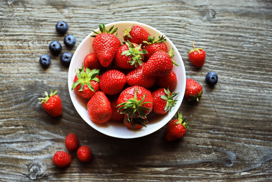 A bowl of fresh strawberries with blueberries and raspberries on a rustic wooden table, shot from above.